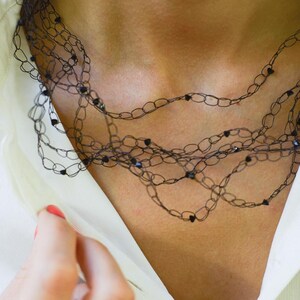 May include: A layered black necklace with small black beads. The necklace is worn over a white collared shirt. The necklace has a delicate, intricate design.
