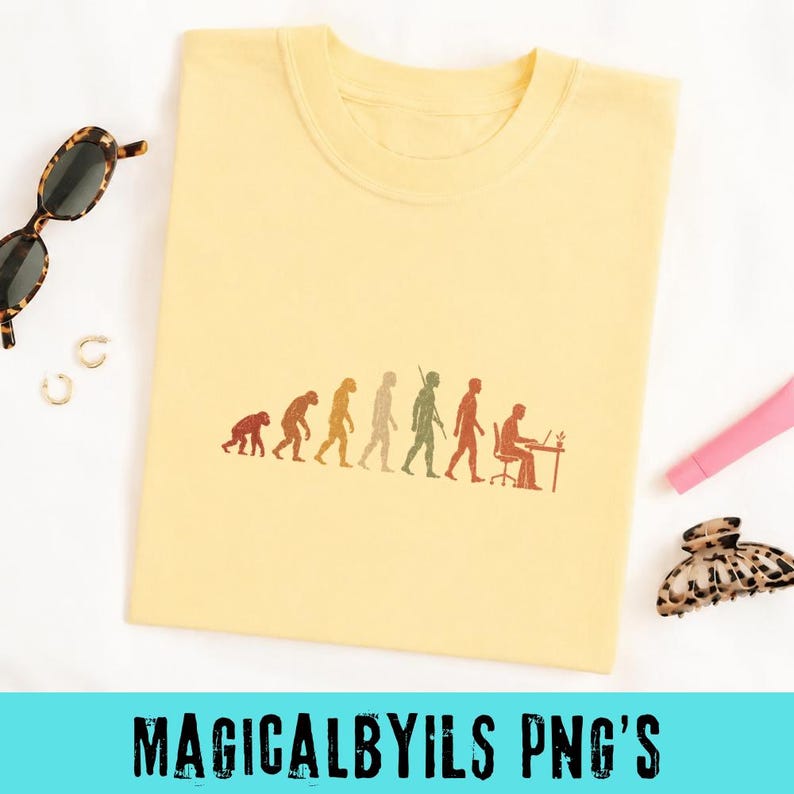 May include: A pale yellow t-shirt featuring a brown graphic print of human evolution, progressing from a primate to a person sitting at a desk with a laptop. The design is centered on the chest of the shirt, which is displayed flat.