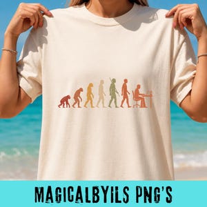 May include: A cream-colored short-sleeve t-shirt featuring a multi-colored graphic print of human evolution. The design shows a progression from a primate to a human sitting at a desk with a computer, rendered in shades of orange, yellow, and green.