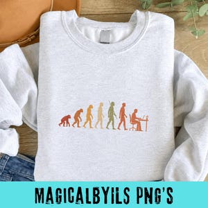 May include: A light gray crewneck sweatshirt featuring a multi-colored graphic print that depicts the evolution of humanity, starting from a primate and progressing through various stages to a person sitting at a desk working on a computer.