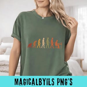 May include: An olive green short-sleeve t-shirt featuring a graphic print of the evolution of humanity, progressing from a primate to a human sitting at a desk with a computer. The design uses a gradient of orange, yellow, and green tones.