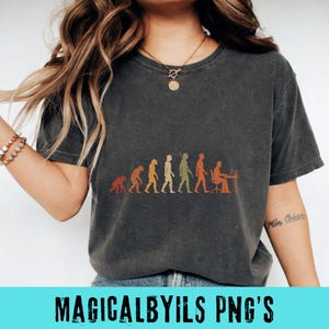May include: A charcoal gray short-sleeve t-shirt featuring a graphic print of human evolution. The design displays a series of figures progressing from a primate to a person sitting at a desk with a computer, rendered in muted orange and green tones.
