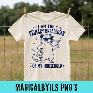 Pode incluir: Uma t-shirt de manga curta cor de creme com um gráfico azul escuro de um gambá a usar óculos de sol, segurando uma mala e um copo de martini. O texto diz: I AM THE PRIMARY BREADLOSER OF MY HOUSEHOLD, rodeado por pequenos detalhes de estrelas.