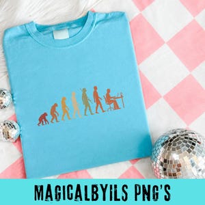 May include: A light blue t-shirt featuring a graphic print of human evolution in shades of orange and yellow, progressing from a primate to a person sitting at a desk with a computer. The design is centered on the front of the crew neck shirt.