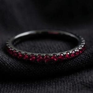 May include: A thin, circular ring featuring a dark, black-toned metal band set with a continuous row of small, vibrant red stones that encircle the entire circumference of the band, creating a striking contrast between the dark metal and red gems.