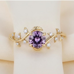 May include: A gold-colored ring featuring a round, purple gemstone at the center. The ring has a leaf and vine design with small, clear accent stones. The ring is set against a cream-colored background.