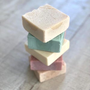 May include: A vertical stack of five rectangular bars of soap. The top bar is off-white with a textured surface, followed by a solid sage green bar, a smooth cream-colored bar, a dusty rose pink bar, and a base bar in a light beige tone.