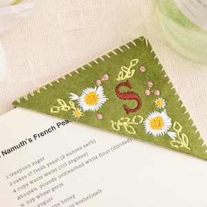 Peut inclure: Marque-page d'angle en feutre vert orné de broderies de marguerites, de feuilles et d'une lettre rouge "S". Le marque-page est posé sur une page de livre de cuisine, avec le titre "Hans Namuth's French Pea".