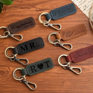 May include: A selection of personalised leather keyrings in shades of brown, black, blue, and burgundy. Each keyring displays engraved text, including names, initials, and phrases like "I Love You" and "To my first hero".