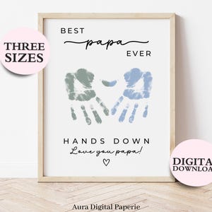 以下が含まれることがあります： 「BEST papa EVER」の文字と、青と緑の手形のデザインが描かれた額入りのプリント。下には「HANDS DOWN Love you papa!」の文字とハートのマーク。プリントは明るい木製のフレームに入っています。