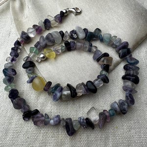 May include: A necklace composed of polished fluorite stones in various colours, including purple, green, yellow, and clear. The necklace features a silver clasp and is presented on a neutral-toned background.