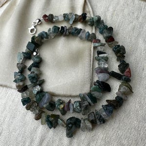 May include: A necklace composed of irregularly shaped, polished green, grey, and red gemstones. The necklace is displayed on cream fabric and has a silver clasp. The gemstones are approximately 1.3 cm in size.