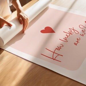 May include: A rolled-up poster with the text "How lucky are we?" in red script, along with a red heart and a playing card symbol. The poster is on a light pink background.