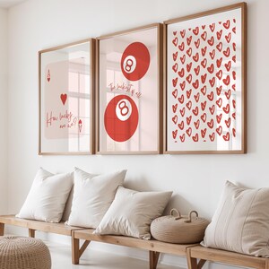 May include: Three framed art prints with wooden frames hang on a white wall. The prints feature a playing card, a red eight ball, and a pattern of red hearts. Below, a wooden bench holds white pillows and a woven basket.