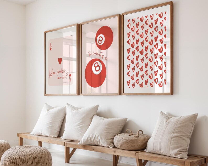May include: Three framed art prints with wooden frames hang on a white wall. The prints feature a playing card, a red eight ball, and a pattern of red hearts. Below, a wooden bench holds white pillows and a woven basket.