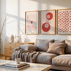 May include: Three framed art prints with a light wood finish hang above a gray sectional sofa. The prints feature a playing card, a pair of red 8-balls, and a pattern of red hearts. A wooden cabinet and decorative items are also visible.