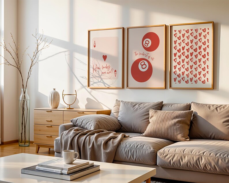 May include: Three framed art prints with a light wood finish hang above a gray sectional sofa. The prints feature a playing card, a pair of red 8-balls, and a pattern of red hearts. A wooden cabinet and decorative items are also visible.