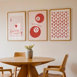 May include: Three framed art prints with a light wood frame hang on a wall. The prints feature a playing card, an eight ball, and a pattern of red hearts. The text "How lucky are we?" is on the playing card print.