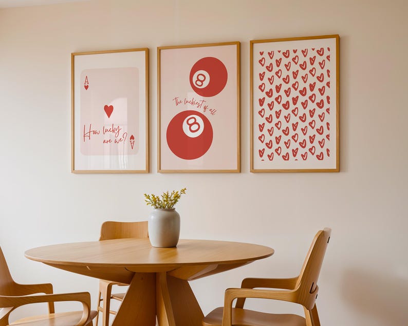 May include: Three framed art prints with a light wood frame hang on a wall. The prints feature a playing card, an eight ball, and a pattern of red hearts. The text "How lucky are we?" is on the playing card print.