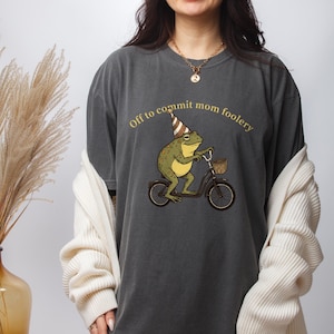 May include: Dark gray t-shirt featuring a cartoon frog riding a bicycle with a basket. The frog wears a party hat and the text "Off to commit mom foolery" is arched above the frog. The shirt is a relaxed fit.