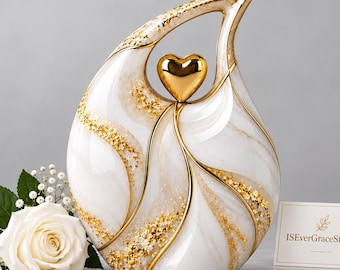 May include: A teardrop-shaped white marble-patterned urn featuring gold leaf accents and a central gold heart. Beside it sits a white rose with baby's breath and a small white card with the text ISEverGraceStudio printed in gold.