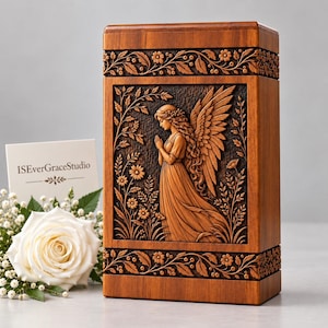 May include: A rectangular wooden urn featuring a carved relief of an angel with wings and long hair standing among flowers. The box has decorative floral borders at the top and bottom. A white card with the text ISEverGraceStudio is visible.