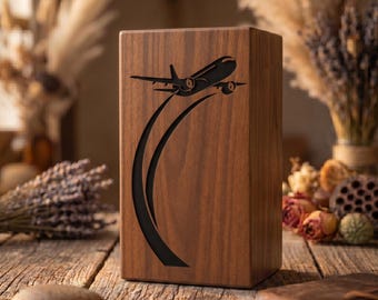 Puede incluir: Una caja de madera rectangular con un acabado marrón oscuro. Un gráfico de avión negro está tallado en la parte delantera, con líneas curvas que lo siguen. La caja se muestra sobre una superficie de madera.