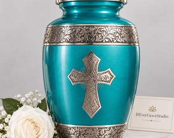 May include: A teal metal cremation urn featuring a silver-toned lid and base with intricate floral engravings. A prominent silver cross with detailed scrollwork is centered on the body. A white rose and a card reading ISEverGraceStudio are present.