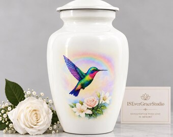 May include: A white cremation urn featuring a colorful hummingbird in flight against a rainbow background, with pink and white flowers below. A small card nearby displays the text ISEverGraceStudio Handcrafted with love in memory.