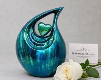 May include: A teardrop-shaped cremation urn with a glossy, metallic teal and blue finish featuring a prominent heart-shaped cutout at the top. A white rose lies in front of the urn next to a white card printed with the text ISEverGraceStudio.