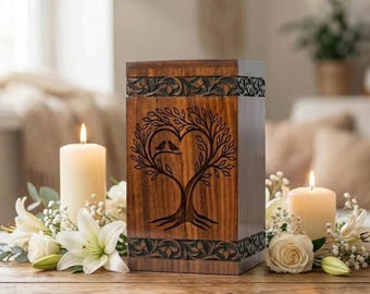 May include: A rectangular wooden cremation urn featuring a dark brown finish with a carved tree of life design forming a heart shape with two birds inside. The top and bottom borders have intricate floral carvings in the wood.