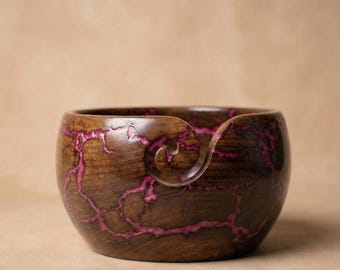 May include: A wooden yarn bowl with a dark brown exterior and vibrant pink veining. The bowl has a smooth, rounded shape with a spiral cutout for yarn. The interior is a darker shade of brown. A decorative and functional item for knitters and crocheters.