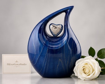 May include: A teardrop-shaped blue cremation urn with a glossy finish and visible wood grain texture. A small, silver-colored heart-shaped emblem is centered in the upper opening. A white card with the text ISEverGraceStudio is placed to the left.