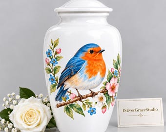 May include: A white ceramic cremation urn featuring a painted illustration of a robin perched on a flowering branch with blue and pink blossoms. The urn has a matching lid and is accompanied by a small white card printed with ISEverGraceStudio.