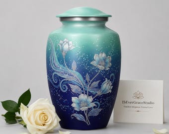 May include: A cremation urn featuring a gradient from light teal at the top to deep navy blue at the base. The surface is decorated with white floral illustrations, swirling lines, and small star-like dots. A white card displays the text ISEverGraceStudio.
