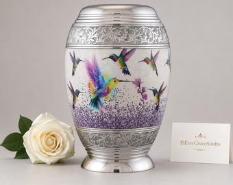 May include: A silver-toned metal cremation urn featuring a colorful hummingbird and lavender floral design. The urn has engraved filigree bands at the top and bottom, a removable lid, and a base. A white rose and a card reading ISEverGraceStudio.