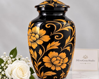 May include: A black cremation urn featuring a vibrant orange floral pattern with swirling vines. The urn has a matching lid and is accompanied by a white rose and a small card with the text ISEverGraceStudio printed in black.