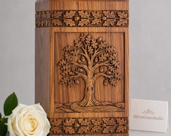 May include: A rectangular wooden urn featuring a detailed relief carving of an oak tree with acorns on the front panel. The top and bottom borders are decorated with a continuous carved oak leaf pattern. A white rose sits beside the urn.