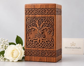 May include: A rectangular wooden cremation urn featuring intricate carvings of a tree and a raven on the front panel. The top and bottom sections are decorated with a continuous Celtic knot pattern. A white rose sits to the left.