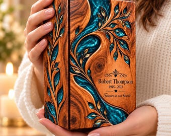 May include: A rectangular wooden cremation urn featuring a swirling teal resin inlay with carved leaf patterns. The front is engraved with the text Robert Thompson, 1948-2023, and Forever in our hearts, accompanied by small heart and scroll motifs.