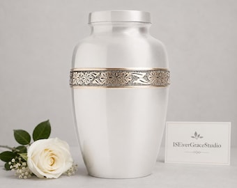 May include: A silver-toned metal cremation urn with a smooth, polished finish and a central horizontal band featuring an embossed floral pattern in a muted bronze color. A white rose with small white filler flowers sits to the left of the urn.