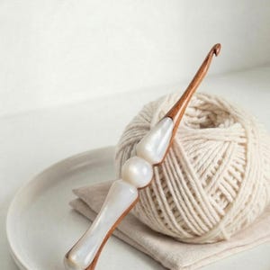 May include: A handcrafted crochet hook with a wooden shaft and a pearlescent white handle. The hook is next to a ball of cream-colored yarn on a white plate and a beige linen napkin.