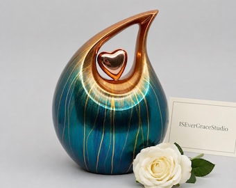 May include: A teardrop-shaped cremation urn featuring a metallic teal and bronze finish with vertical gold-toned streaks. A small, polished bronze heart is suspended within the upper opening. A white rose and a card reading ISEverGraceStudio.