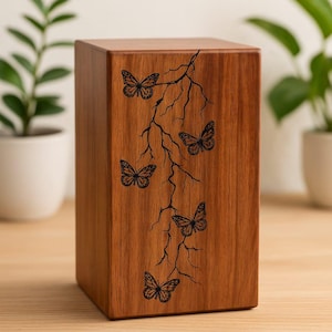 Puede incluir: Una caja de madera con acabado natural, con un dise&ntilde;o tallado de mariposas y ramas. La caja es rectangular y se alza verticalmente, con el dise&ntilde;o de mariposa en vertical. El fondo est&aacute; borroso, con plantas visibles.