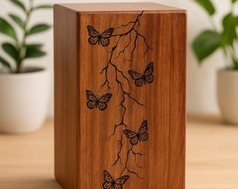 May include: A wooden box with a natural finish, featuring a carved design of butterflies and branches. The box is rectangular and stands upright, with the butterfly design running vertically. The background is blurred, with plants visible.