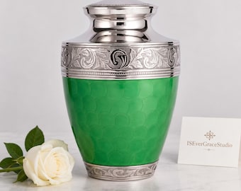May include: A metal cremation urn featuring a vibrant green body with a scalloped pattern, topped with a silver lid and a silver base. The upper section is adorned with intricate silver floral engravings. A white rose sits beside the urn.