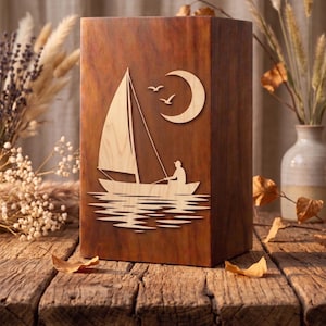 Puede incluir: Caja de madera con diseño de velero. El velero está representado con una persona en el barco, navegando sobre el agua, con una luna y pájaros en el cielo. La caja es de madera marrón oscuro y está sobre una superficie de madera.