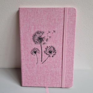Può includere: Diario rosa con un design a soffione. La copertina ha un aspetto strutturato e presenta un'illustrazione nera di capolini di soffione. Sono inclusi un elastico e un segnalibro abbinati.