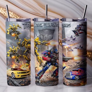 May include: Three cylindrical tumblers with metal straws featuring Transformers graphics. The designs include yellow and blue robot characters, a yellow sports car, a blue truck, fighter jets, and the Transformers logo printed in silver metallic lettering.