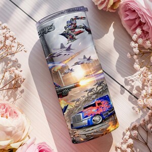 May include: A cylindrical metal tumbler featuring a full-wrap graphic print of a battle scene with a blue and red semi-truck, fighter jets, and a robotic character. The design includes fiery explosions and the partial text ERS in a metallic font.